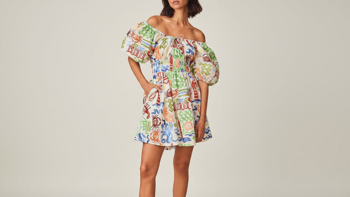 An off-the-shoulder mini dress with a colorful, busy illustrated print, worn by a model in green strappy heels against a solid beige studio background.